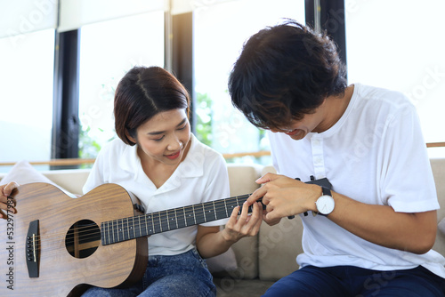 An asian man teaching a woman how to play acoustic guitar, by pointing out the placement of fingers on the fingerboard as a weekend family activity. Asian couple concept.