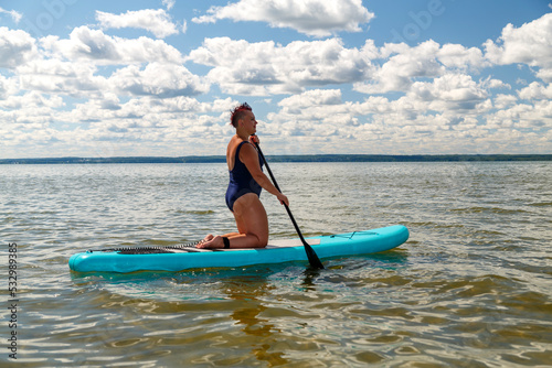 a woman in a closed swimsuit with a mohawk on her knees on a SUP board with an oar floats on the water. © finist_4