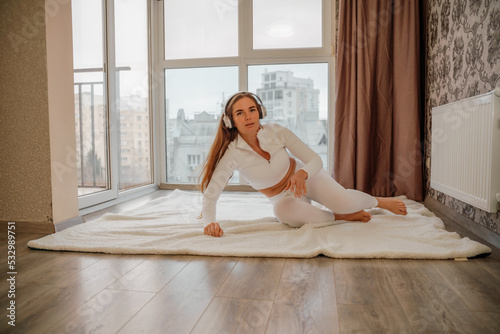 Side view portrait of relaxed woman listening to music with headphones lying on carpet at home. She is dressed in a white tracksuit.