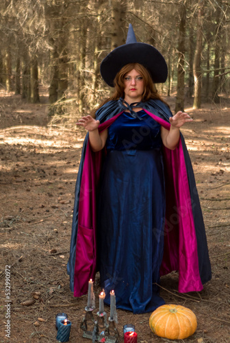 a witch in a blue cloak tells fortunes in the forest, makes movements with her hands and reads spells next to a candle and a pumpkin.