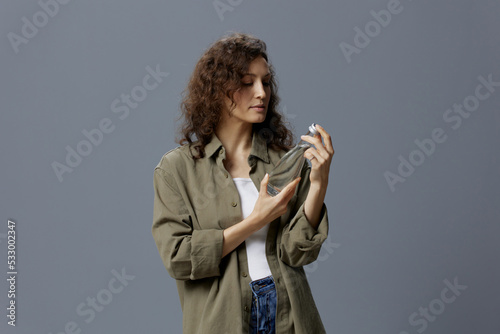 Serious focused curly beautiful woman in casual khaki shirt hold open water bottle maintains water balance posing isolated on gray background. Healthy lifestyle. Water is Life concept. Copy space