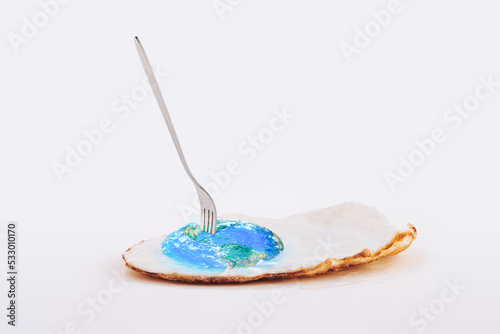 Planet Earth pierced with fork as a part of fried egg on isolated pastel white background. Minimal abstract concept of dystopian future, global warming or climate changing. World provided by NASA.