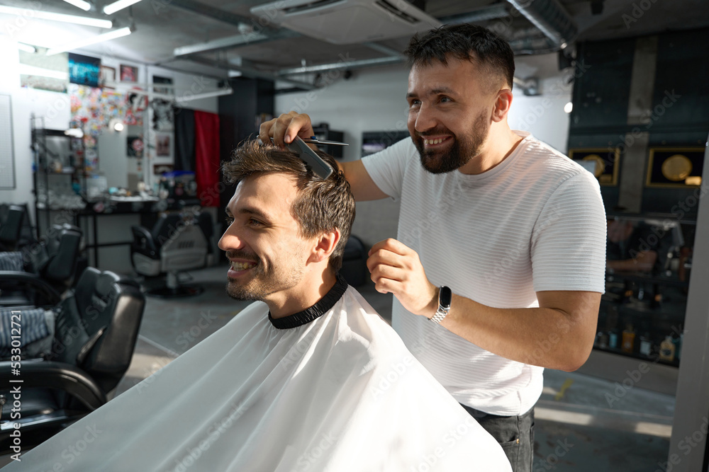 Obraz premium Cheerful barbershop worker smiling while combing young brunet guy hair