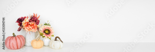 Autumn decor on a white shelf against a white wall banner background. Pumpkins and flowers in vases with pink hue fall colors. Copy space.