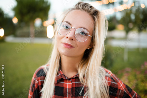 Smiling beautiful young adult student female girl wear glasses over glow lights and nature green background outdoor close up. Look at camera. Back to school.