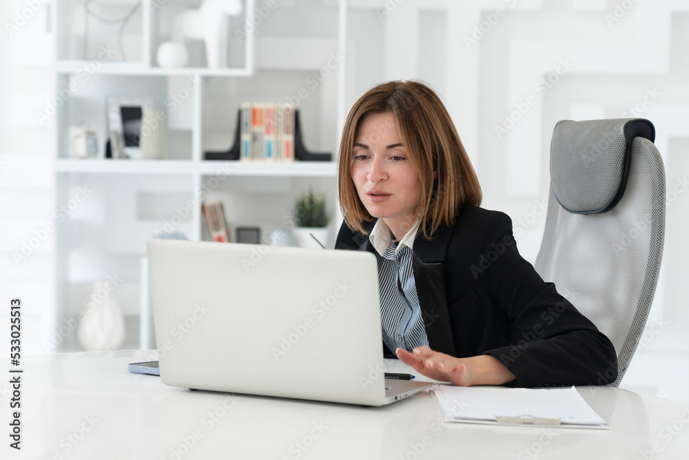 Portrait of successful and attractive business woman working on laptop in her office. Positive female employee in black suit smiling and sitting at the desk in her modern workplace Portrait of successful and attractive business woman working on laptop in her office. Positive female employee in black suit smiling and sitting at the desk in her modern workplace