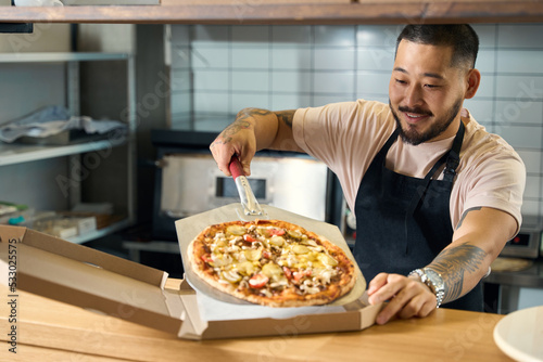 Pleasant young man in cook apron packing pizza into box