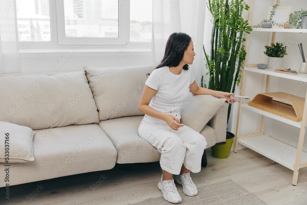 A young Asian woman sits at home on the couch relaxing and flipping through her social media feed and reading messages on her phone blogger home clothes. Lifestyle without work in a comfortable home