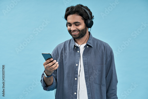 Happy smiling young arab ethnic man wearing headphones using smartphone looking at cell phone watching videos, having virtual mobile chat video call, playing game isolated blue background.