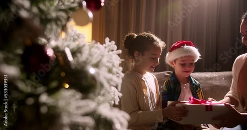 Happy family with little daughter and son in Santa hats gathered together on Christmas morning near xmas tree chatting enjoy gifts opening. New Year celebration, presents store advertisement concept