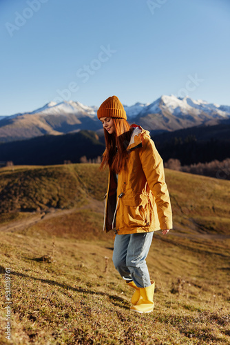 Full-length woman running up the hill to the camera and looking at the mountains in a yellow raincoat and jeans in the autumn happy sunset hiking trip mountains in the snow, freedom lifestyle 