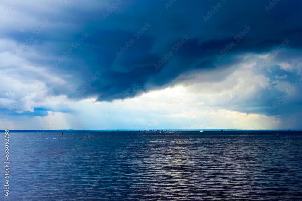 Sea, dark clouds in the sky, huge number of sailboats on the sea in the sunlit area. Beautiful colorful sky with an approaching storm
