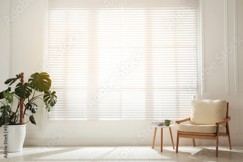 Obraz na plátne Cosy armchair and houseplant near large window with blinds in spacious room