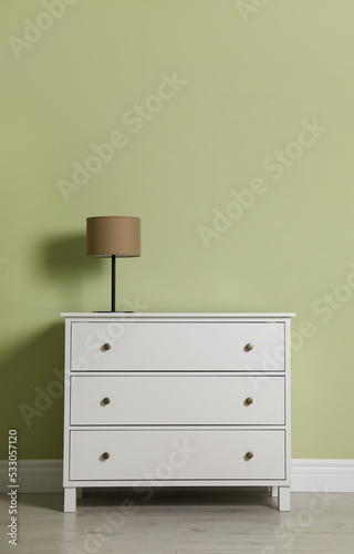 Modern white chest of drawers with lamp near light green wall indoors © New Africa
