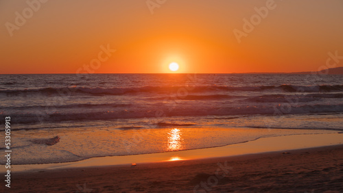 Sunset seen at Santa Monica Beach, CA, USA in Summer 2022. Beautiful orange tones in the twilight sky. Sun in perfect circle sets into the Pacific Ocean.