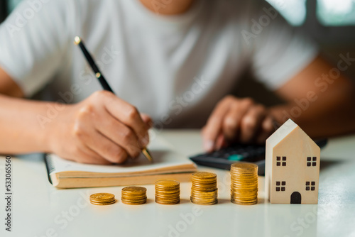 Businessman using calculator to calculate and taking notes and Stacking coins, wooden house on table. Saving money,accounting, investment, budgeting, and financial planning concept.