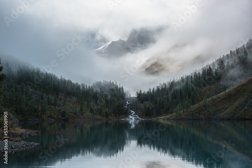 Mountain creek flows from forest hills into glacial lake. Snow rock mountains in fog clearance. Small river and coniferous trees reflected in calm alpine lake. Tranquil misty scenery at early morning.