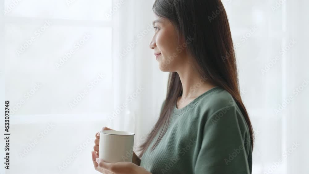 Young asian woman standing beside window and holding mug in bedroom at home, She drinking milk after wake up in the morning