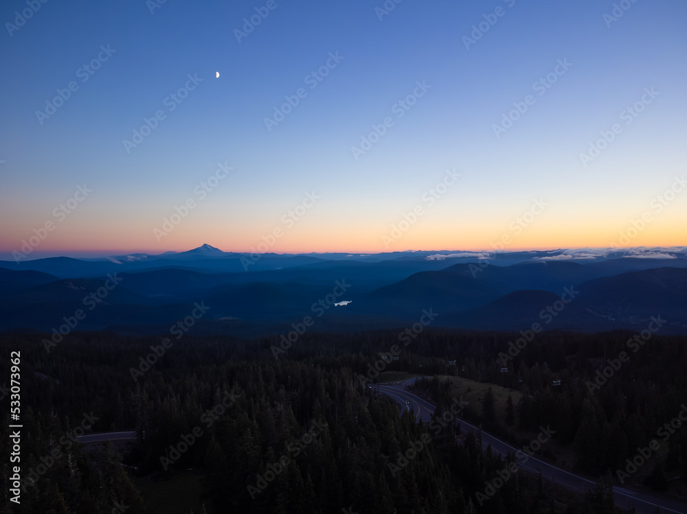 Shooting from the air. Stunning night landscape. Automobile route among the forest and mountains. The sky is colored with orange rays of the setting sun. Tourism, travel, nature.