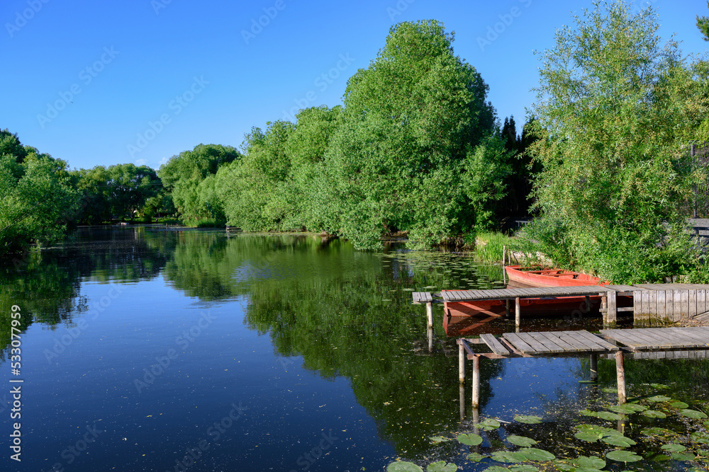 A river with water lilies, wooden piers and a boat surrounded by trees in summer