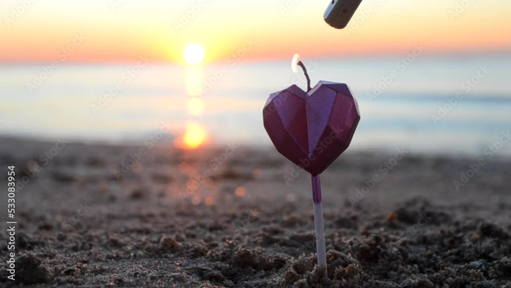 A man lights a candle in the shape of a heart on the sea sandy beach at dawn sunset. Candle in shape of heart burning on background of sea waves on dawning close-up. Romantic mood. Valentine's Day A man lights a candle in the shape of a heart on the sea sandy beach at dawn sunset. Candle in shape of heart burning on background of sea waves on dawning close-up. Romantic mood. Valentine's Day