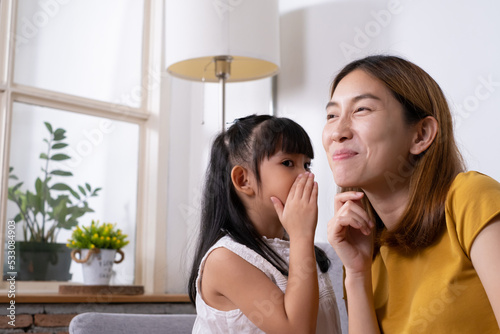 Asian mother feeling surprise and happy with smile while her daughter tell or whispering news and story to her inside of living room. Mom and young girl relationship, parenthood.