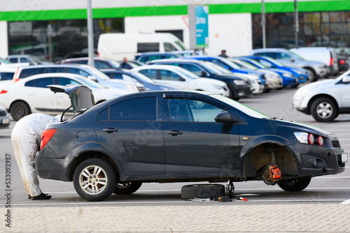 A car with a wheel removed is on a jack in a supermarket parking lot. The male driver takes a tool out of the trunk to fix a flat tire.