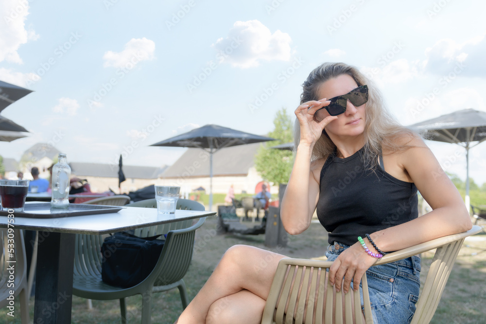 Young beautiful woman drinking water, in an outdoor cafe, enjoying a sunny morning. Looking away and touching her sunglasses. In a stylish black blouse and denim short skirt