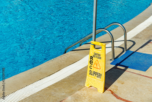 Safety sign with phrase Caution wet floor near the swimming pool at the tropical resort. Cleaning service slippery