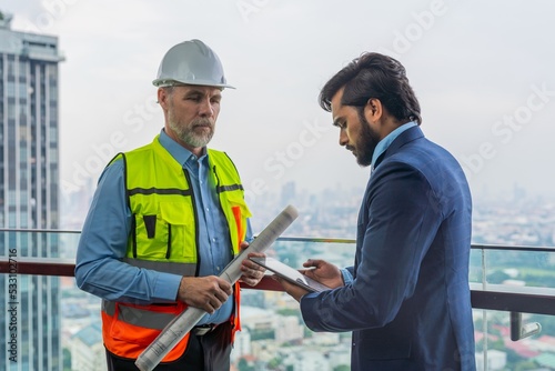 Portrait photo of a white caucasian construction architect holding a architecture plan discussing the project details with his client an asian businessman holding a digital tablet