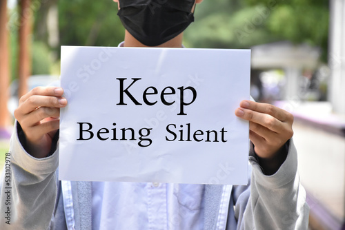 Asian boy student holds white paper which has texts 'Keep Being Silent' inside, concept for protesting or calling out all people around the world to keep being silent or keep quiet.