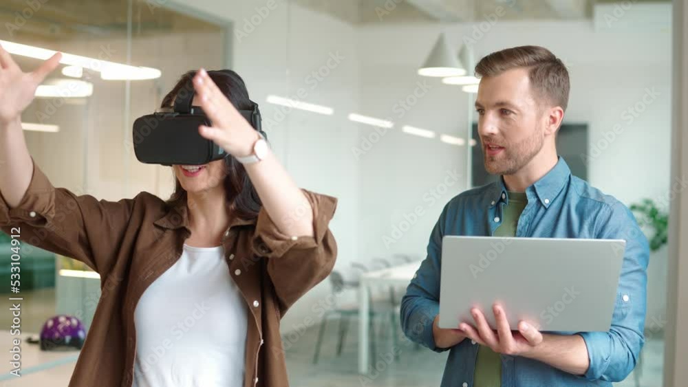 Close up portrait of handsome young male manager holding laptop and speaking while woman wearing VR headset and moving hands in air. Virtual reality presentation. Office work. Futuristic technology Close up portrait of handsome young male manager holding laptop and speaking while woman wearing VR headset and moving hands in air. Virtual reality presentation. Office work. Futuristic technology