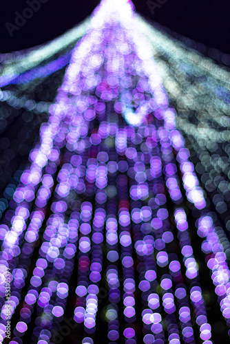Festive purple xmas lights on christmas tree in urban central park at holiday winter night, defocus shiny garland decoration glow in lilac bokeh in dark downtown, vertical out of focus image