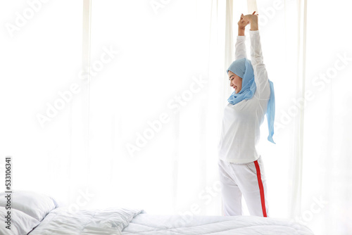 Beautiful asian woman wearing white muslim sleepwear, stretching her arms after getting up in the morning at sunrise. Cute young girl with blue hijab standing and relaxing with happy and smiling face. © feeling lucky