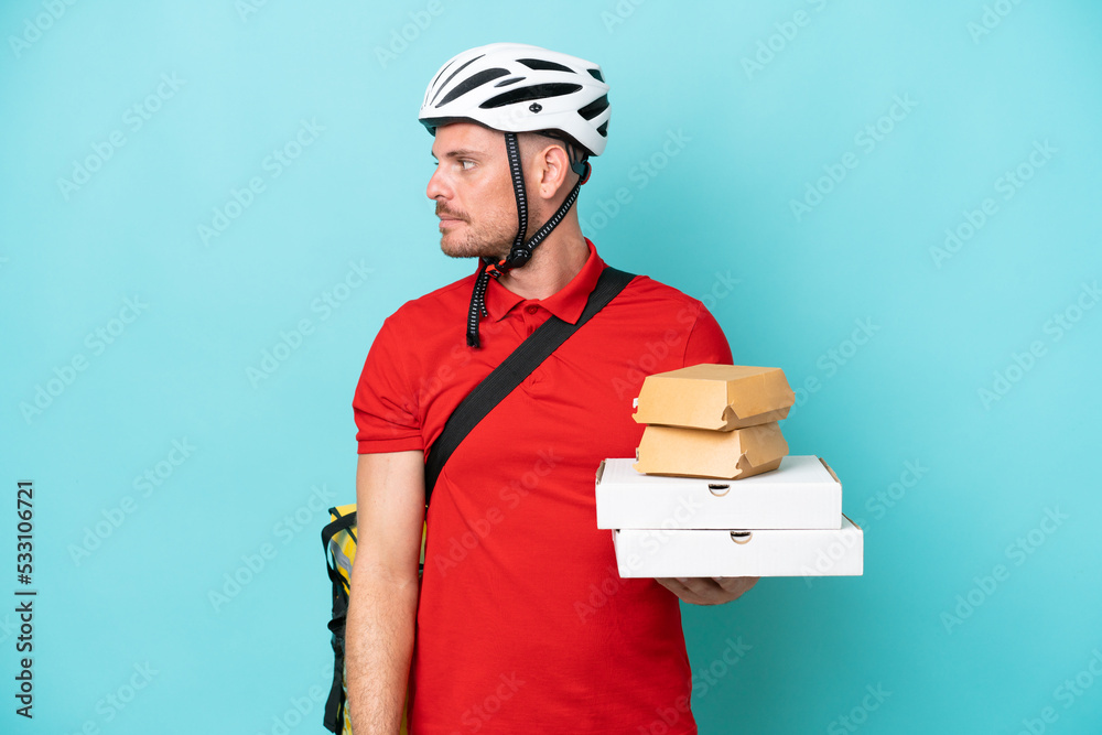Young caucasian man with thermal backpack and holding fast food isolated on blue background looking to the side Young caucasian man with thermal backpack and holding fast food isolated on blue background looking to the side