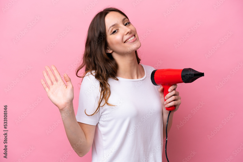 Young caucasian woman holding a hairdryer isolated on pink background saluting with hand with happy expression Young caucasian woman holding a hairdryer isolated on pink background saluting with hand with happy expression