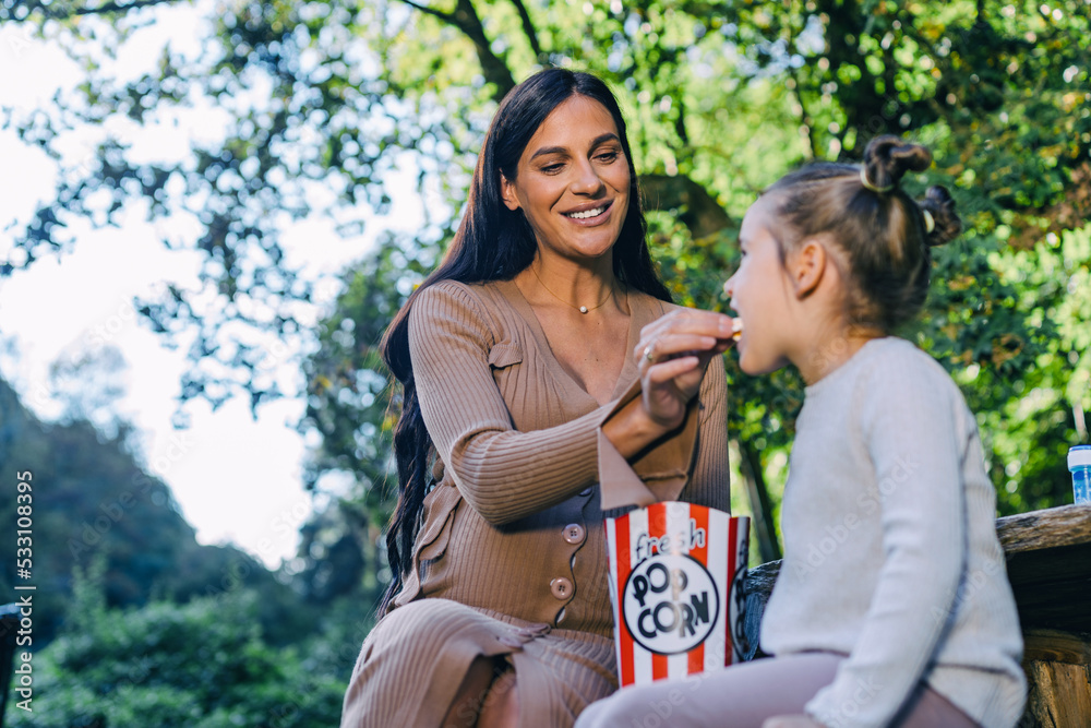 A pregnant woman and her daughter are sitting on a bench in an autumn park and eating popcorn, having fun together. A pregnant woman and her daughter are sitting on a bench in an autumn park and eating popcorn, having fun together.