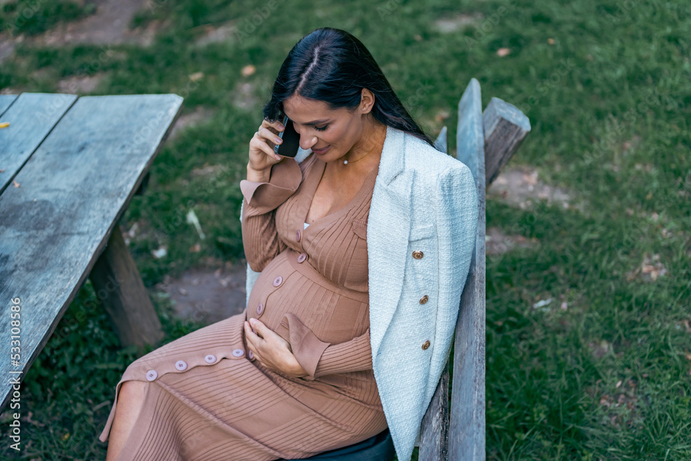 Naklejka premium Pregnant woman sits on bench in the park and using phone. Happy female have rest outdoor during the walks