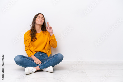 Young caucasian woman sitting on the floor isolated on white wall with fingers crossing and wishing the best