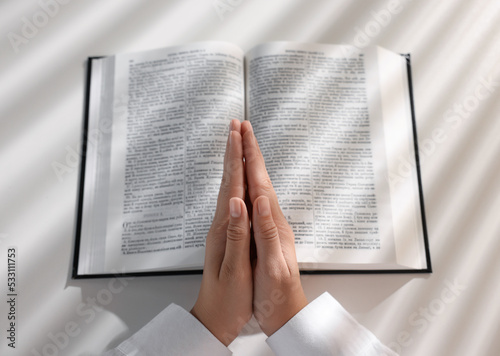 Above view of woman holding hands clasped while praying over Bible at white table