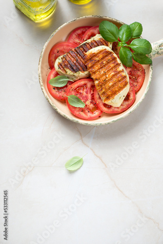 Serving pan with grilled halloumi, red tomatoes and green basil, above view on a beige stone background, vertical shot, copy space