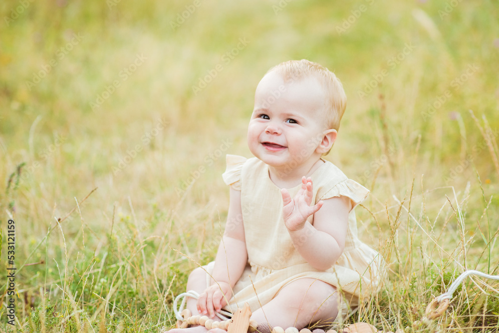 Happy baby girl waving hello sitting on yellow grass lawn. Kid sitting in autumn park Happy baby girl waving hello sitting on yellow grass lawn. Kid sitting in autumn park