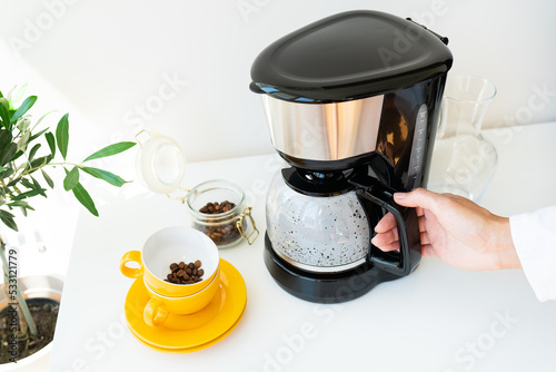 Beautiful morning, the girl is waiting for her coffee. An automatic drip coffee maker stands with a yellow cup on a white table. Electric kitchen small household appliances. photo