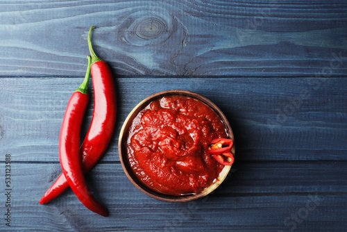 Composition of hot tomato sauce in a wooden bowl on a blue wooden background. Tomato sauce in a wooden bowl, slices of fresh chili peppers, A branch of ripe cherry tomatoes, Fresh allspice basil.