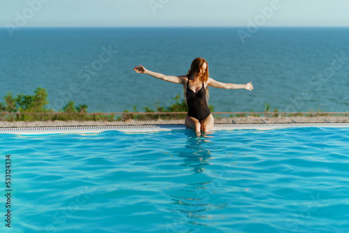 A slender woman in a black swimsuit sits on the side of a panoramic pool. 