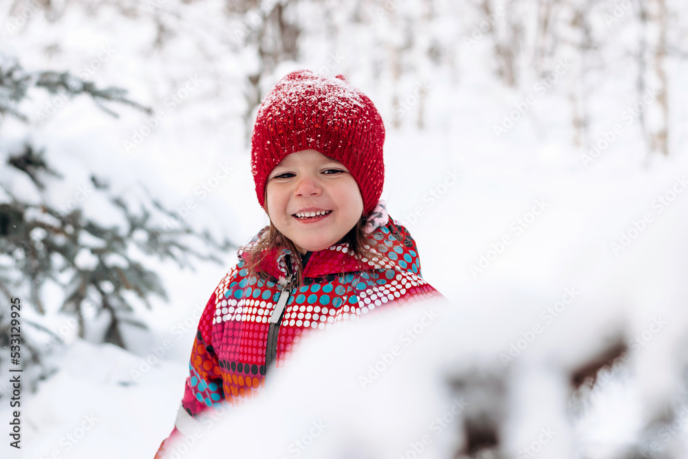 Happy little girl in a red hat and jumpsuit walks in the winter forest.Beautiful trees are covered with white snow.Winter fun,active lifestyle concept.