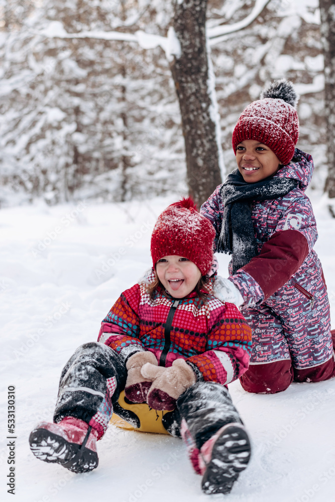 Happy Caucasian and African-American girls ride a saucer in the winter park.Beautiful trees are covered with white snow.Winter fun,active lifestyle concept.