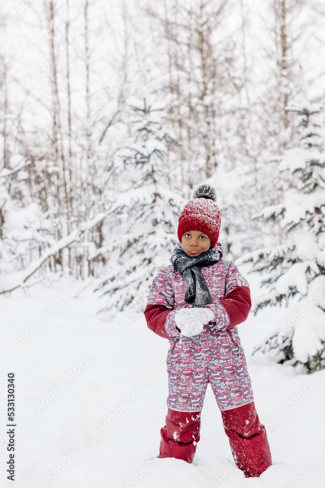 Happy little African-American girl in red hat and jumpsuit walks in the winter forest and throws up snow.Beautiful trees are covered with white snow.Winter fun,active lifestyle concept.Selective focus