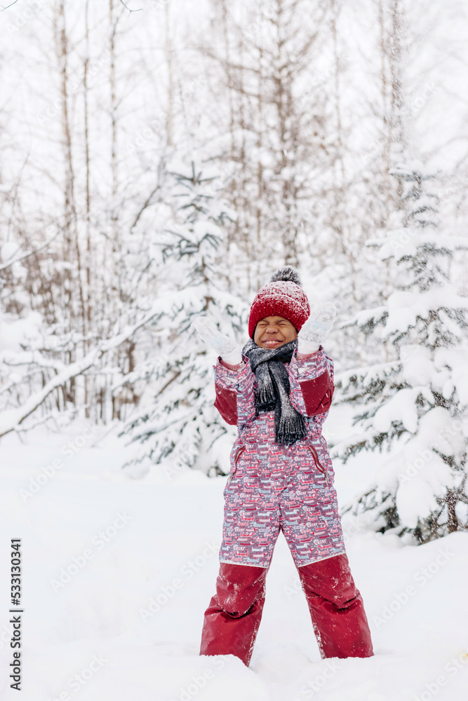 Happy little African-American girl in red hat and jumpsuit walks in the winter forest and throws up snow.Beautiful trees are covered with white snow.Winter fun,active lifestyle concept.Selective focus
