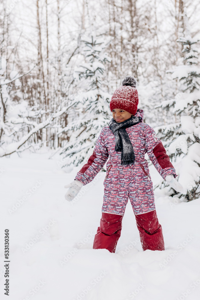 Happy little African-American girl in red hat and jumpsuit walks in the winter forest and throws up snow.Beautiful trees are covered with white snow.Winter fun,active lifestyle concept.Selective focus